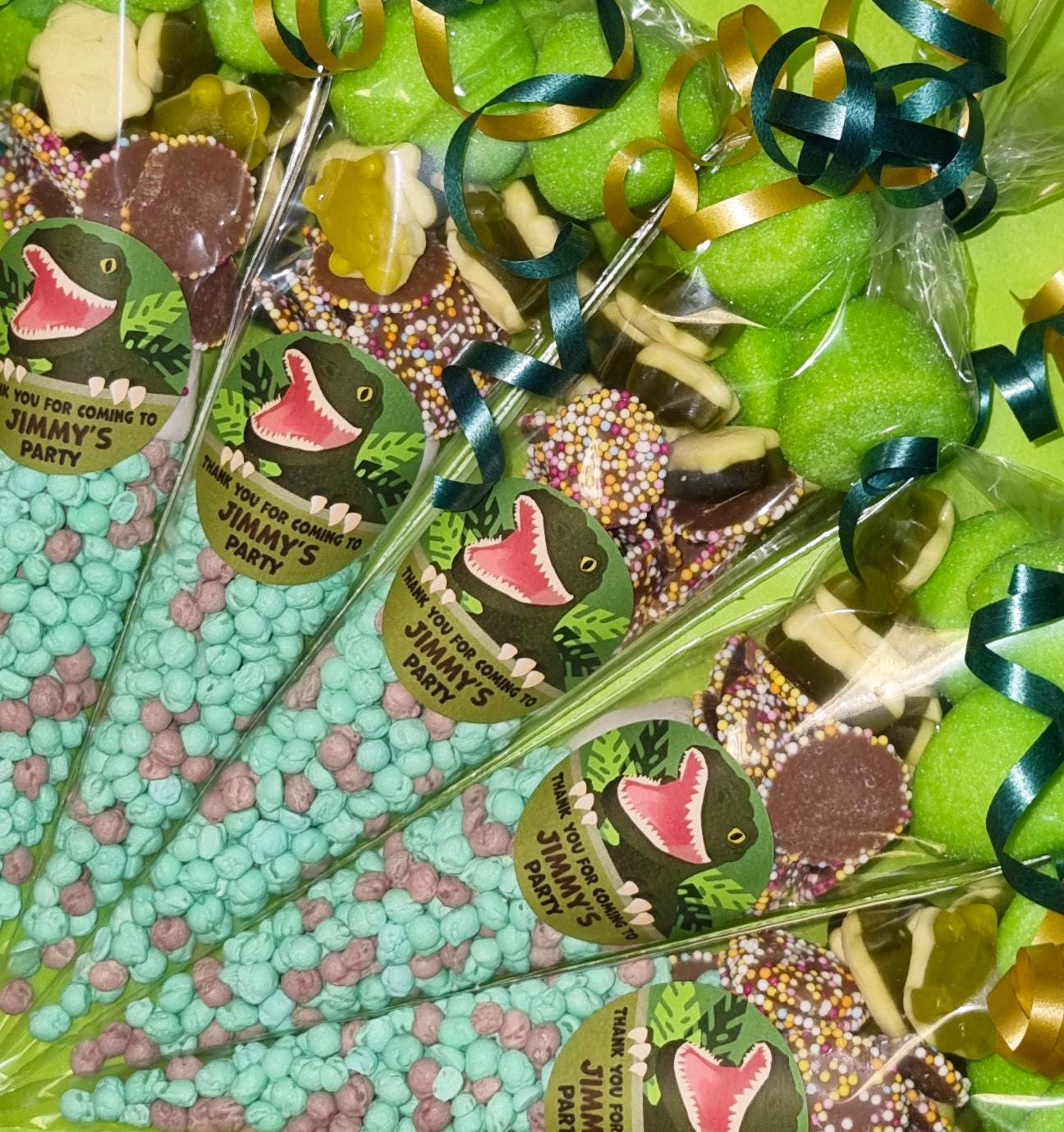 Pre-filled sweet cones with dinosaur motifs and various candies visible inside, wrapped in clear plastic with green and blue ribbons.