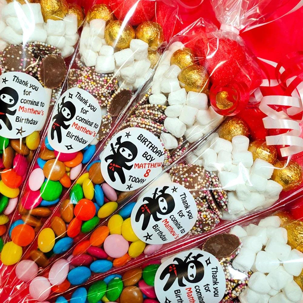 Pre-filled sweet cones with a Ninja Party theme, containing various candies and chocolates, wrapped in transparent packaging with red decorative elements and labels with character illustrations and text.